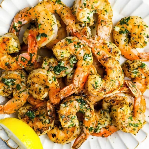 Grilled shrimp with herbs on a white scalloped plate, accompanied by a lemon wedge. The shrimp are seasoned with herbs and appear golden and slightly charred.