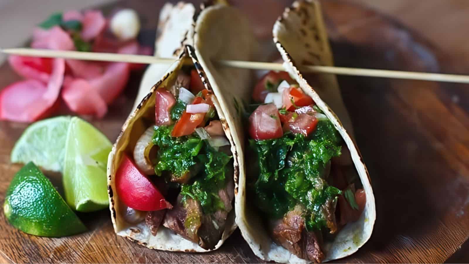 Two tacos filled with grilled meat, diced tomatoes, chopped onions, and green salsa, garnished with a lime wedge on the side and pickled onions in the background on a wooden surface.