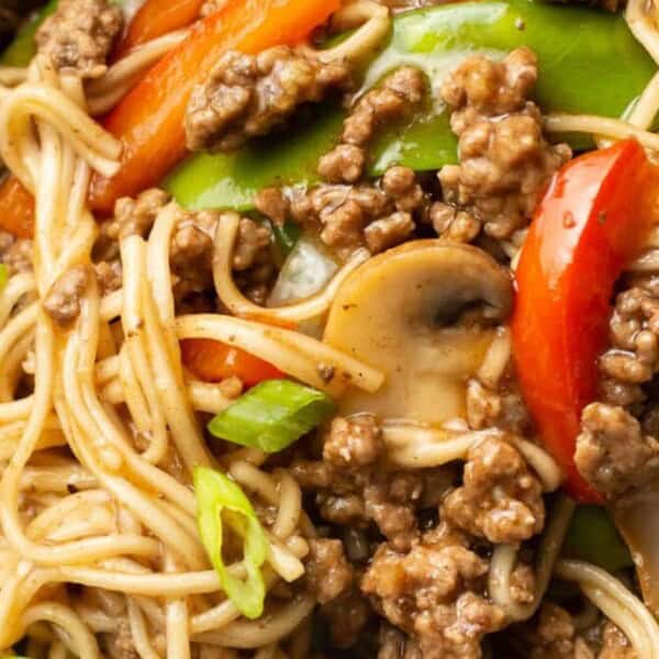 A close-up of a dish featuring noodles mixed with ground beef, sliced mushrooms, red bell peppers, and snow peas. Green onions are garnished on top, adding a splash of color and freshness.