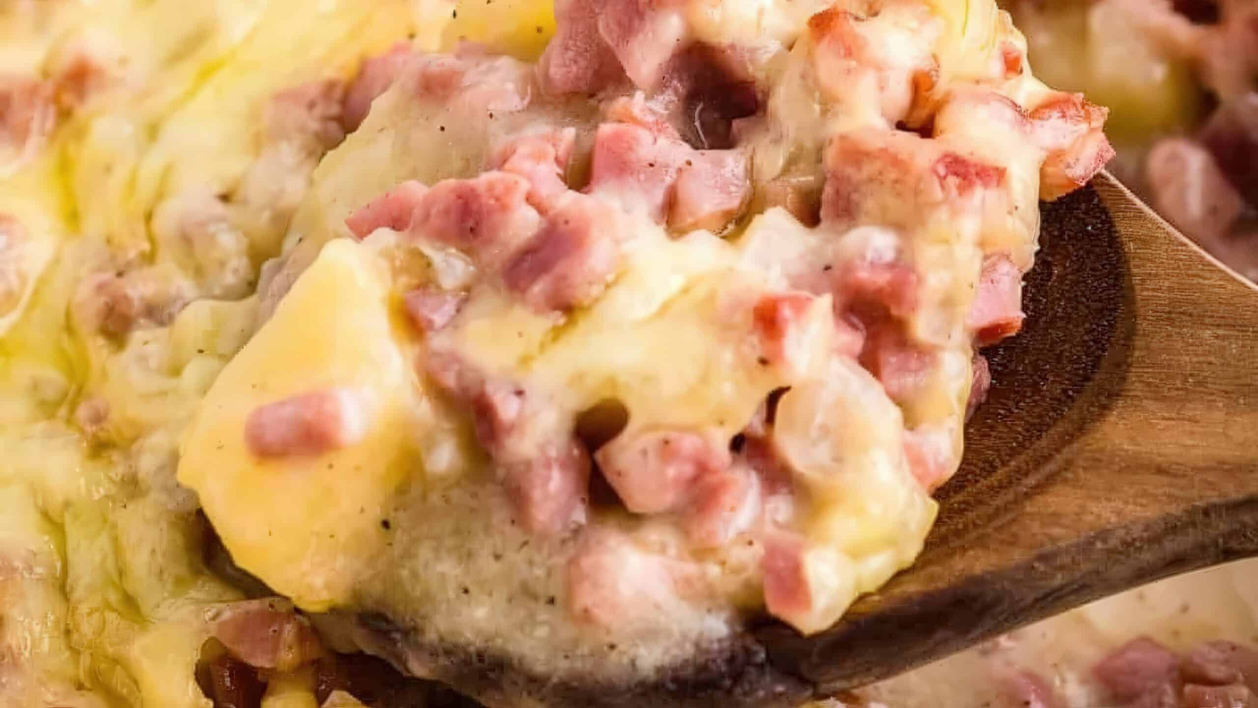 A close-up of a wooden spoon scooping a creamy mixture of scrambled eggs, melted cheese, and diced ham. The dish appears rich and savory, with a mix of soft textures and a slightly golden hue.