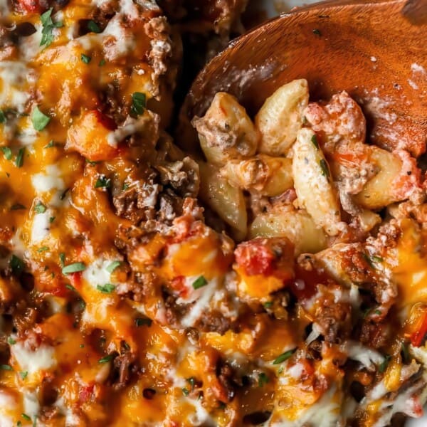 Baked pasta casserole with melted cheese, ground beef, and tomatoes, served from a white dish with a wooden spoon.