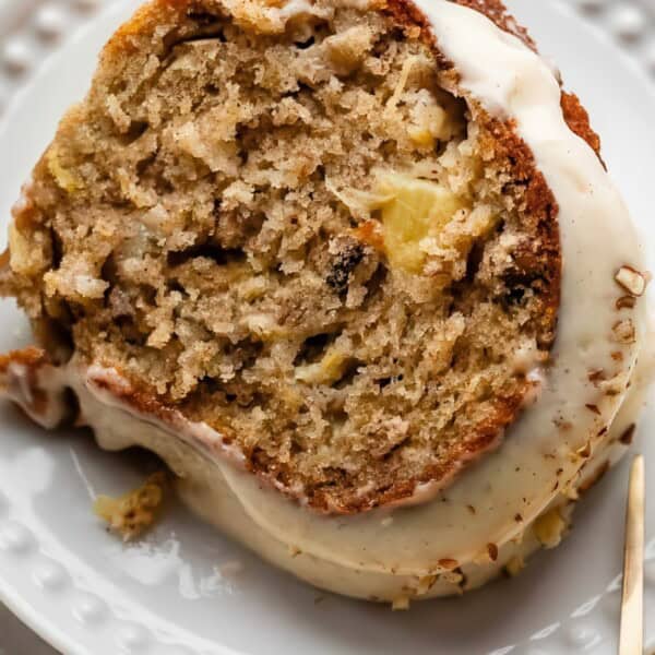 A slice of moist cake with chunks of fruit and nuts, topped with a white frosting on a white plate. Nearby, a beige napkin and a fork rest on the table.