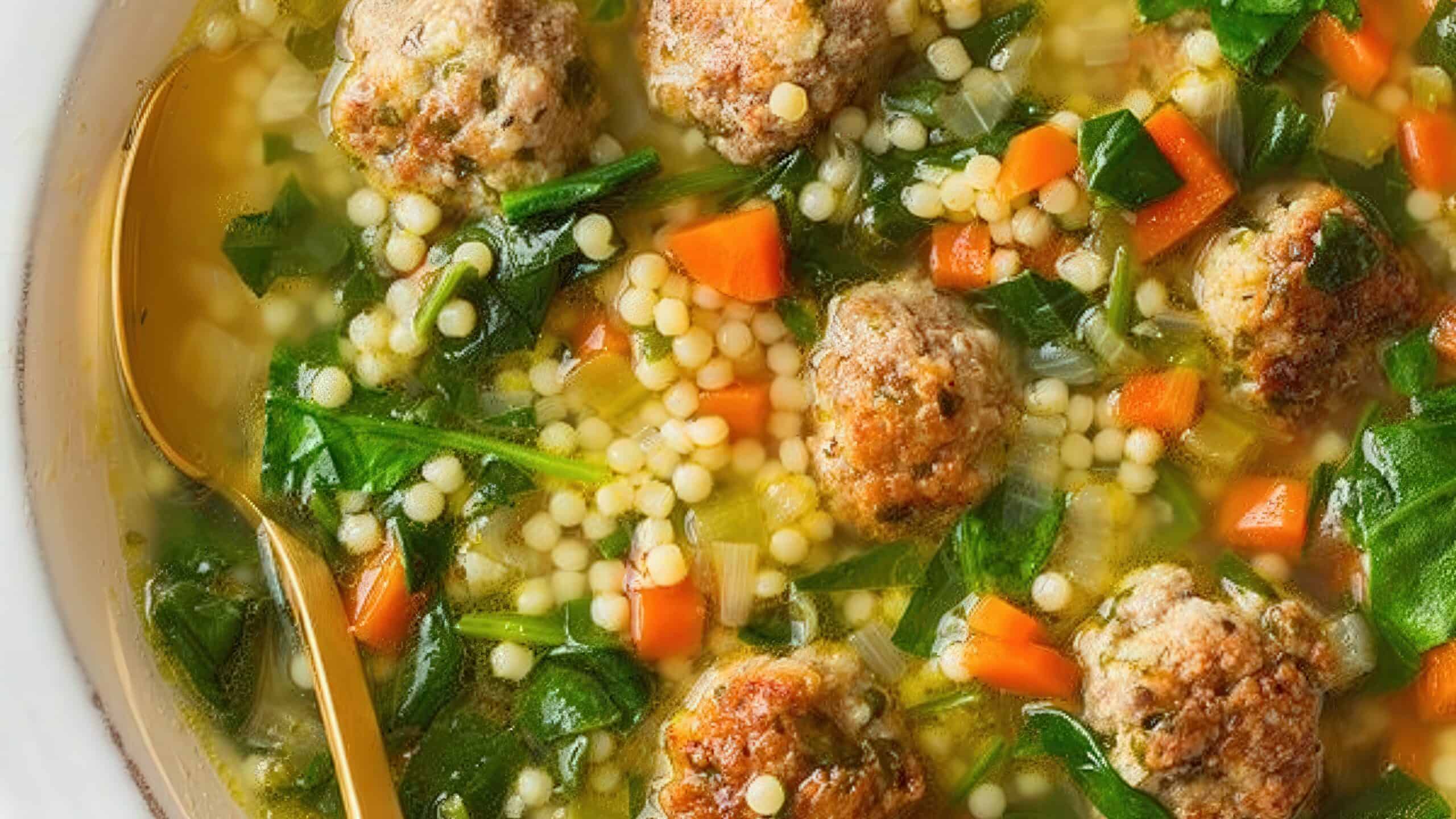 A close-up of a bowl of Italian wedding soup, featuring meatballs, small pasta, spinach, and diced carrots in a clear broth. A gold spoon is partially visible on the side of the bowl.
