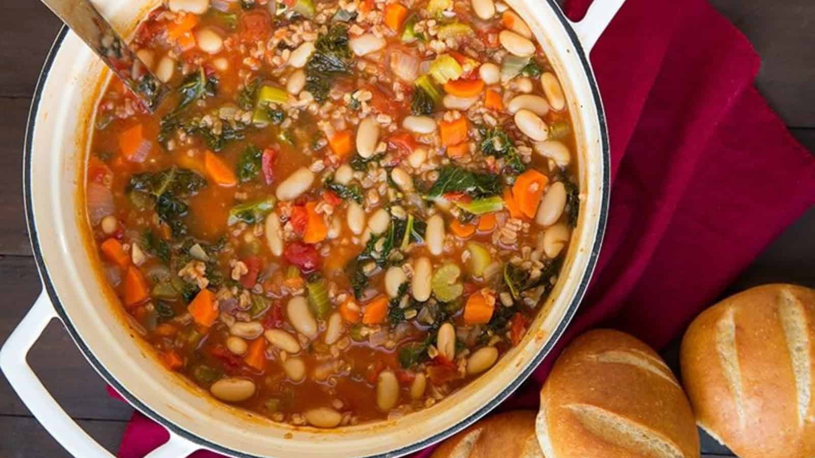 A pot of hearty vegetable soup with beans, carrots, tomatoes, and greens sits on a dark wooden table. Next to it are round loaves of bread on a red cloth. A ladle rests inside the pot.