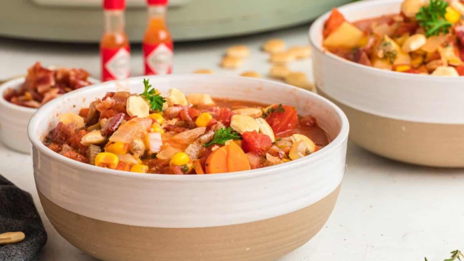 Two bowls of hearty soup filled with vegetables like tomatoes, corn, and carrots, topped with parsley and crackers. Bottles of hot sauce are visible in the background. The setting is bright and inviting.