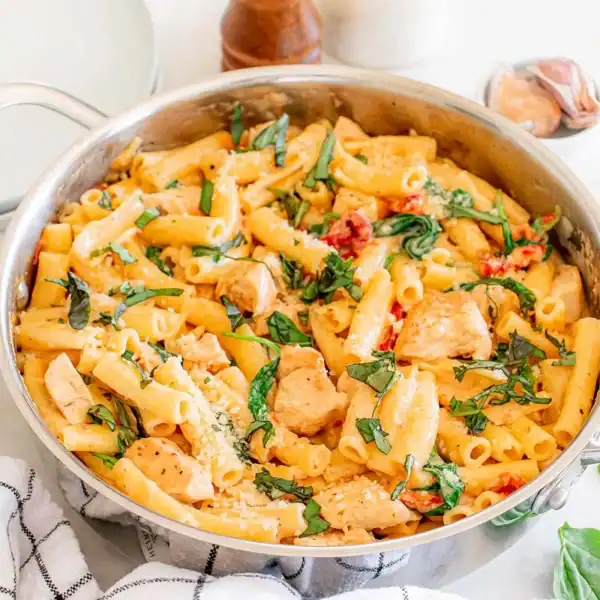 A stainless steel pan with creamy rigatoni pasta, chicken pieces, spinach, sun-dried tomatoes, and grated cheese on a white surface.