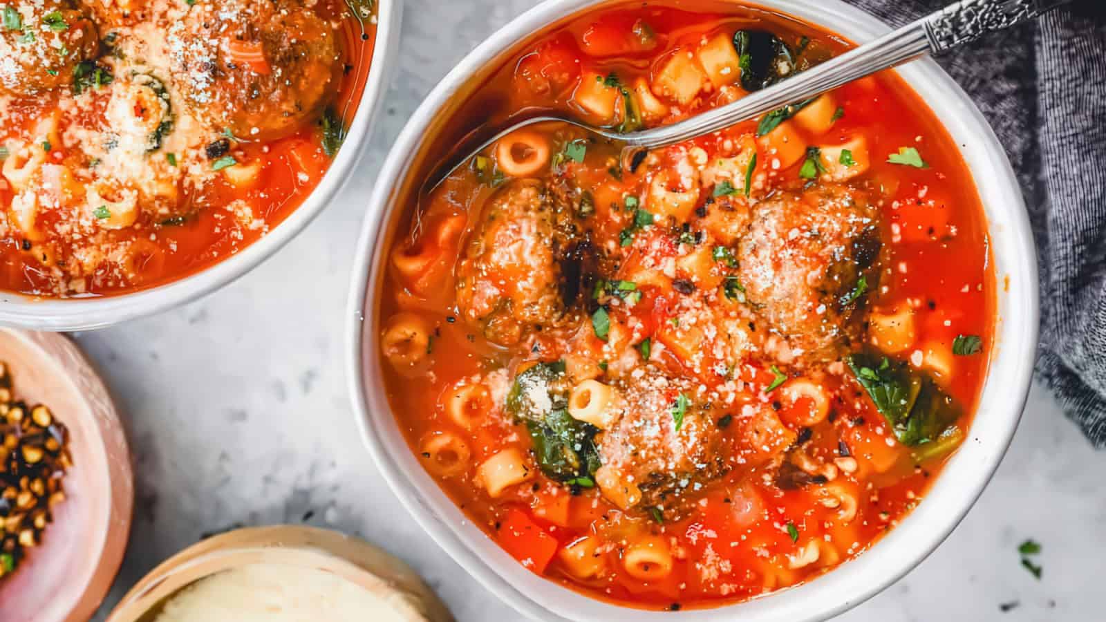 A bowl of hearty tomato soup with pasta and meatballs, garnished with grated cheese and fresh herbs. A spoon rests in the bowl, surrounded by more bowls and a sprinkle of crushed red pepper on the side.