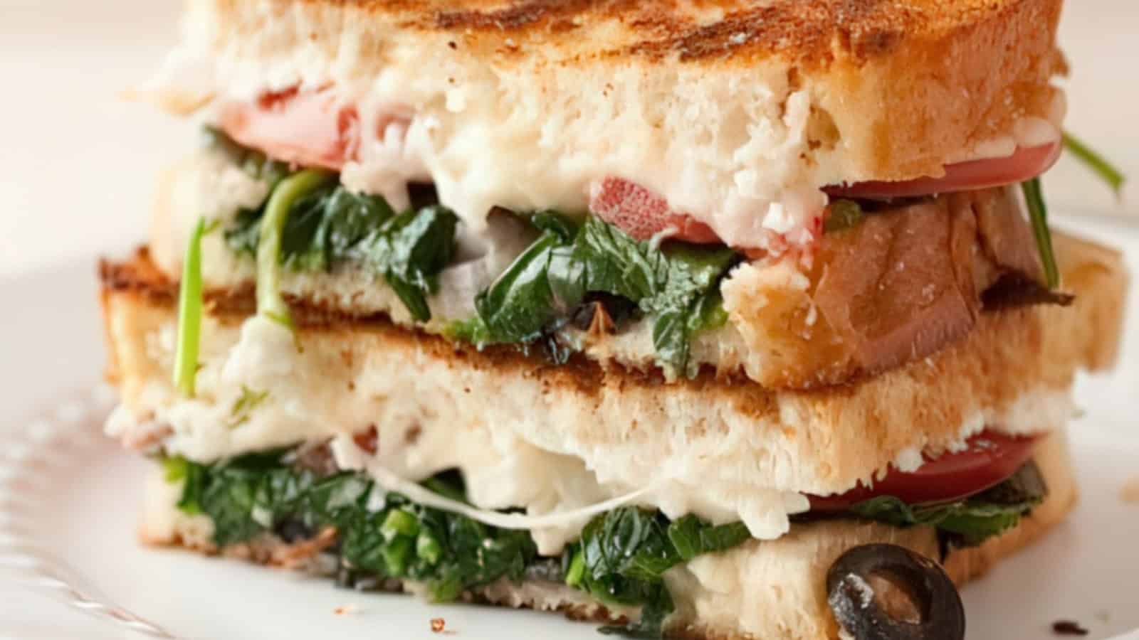 A stacked sandwich with toasted bread, filled with melted cheese, sliced tomatoes, fresh spinach, and black olives. The sandwich is cut in half and placed on a white plate.
