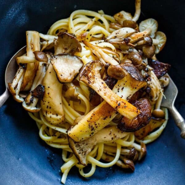 A bowl of pasta topped with an assortment of sautéed mushrooms, including shiitake and oyster varieties. Two serving utensils rest on either side of the bowl. The dish is served in a dark blue bowl.