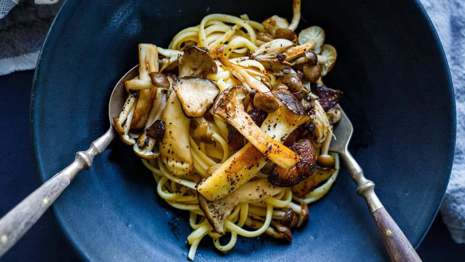 A bowl of pasta topped with an assortment of sautéed mushrooms, including shiitake and oyster varieties. Two serving utensils rest on either side of the bowl. The dish is served in a dark blue bowl.