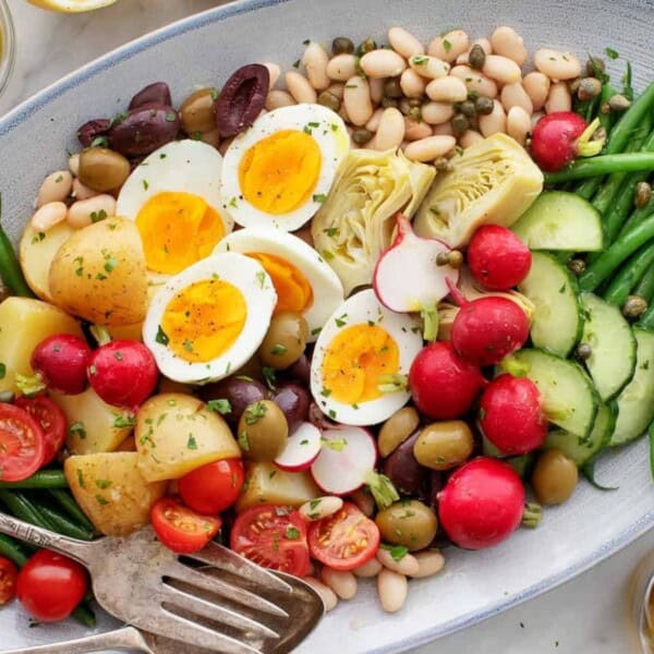 An oval platter featuring a colorful salad with hard-boiled eggs, cherry tomatoes, radishes, cucumbers, olives, green beans, potatoes, white beans, and artichoke hearts, garnished with herbs. A serving fork is on the side.