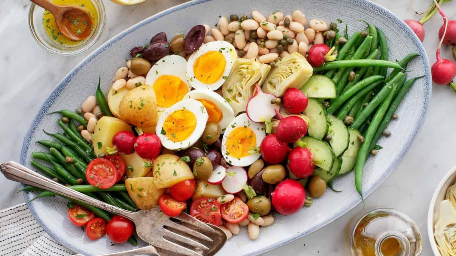 An oval platter featuring a colorful salad with hard-boiled eggs, cherry tomatoes, radishes, cucumbers, olives, green beans, potatoes, white beans, and artichoke hearts, garnished with herbs. A serving fork is on the side.