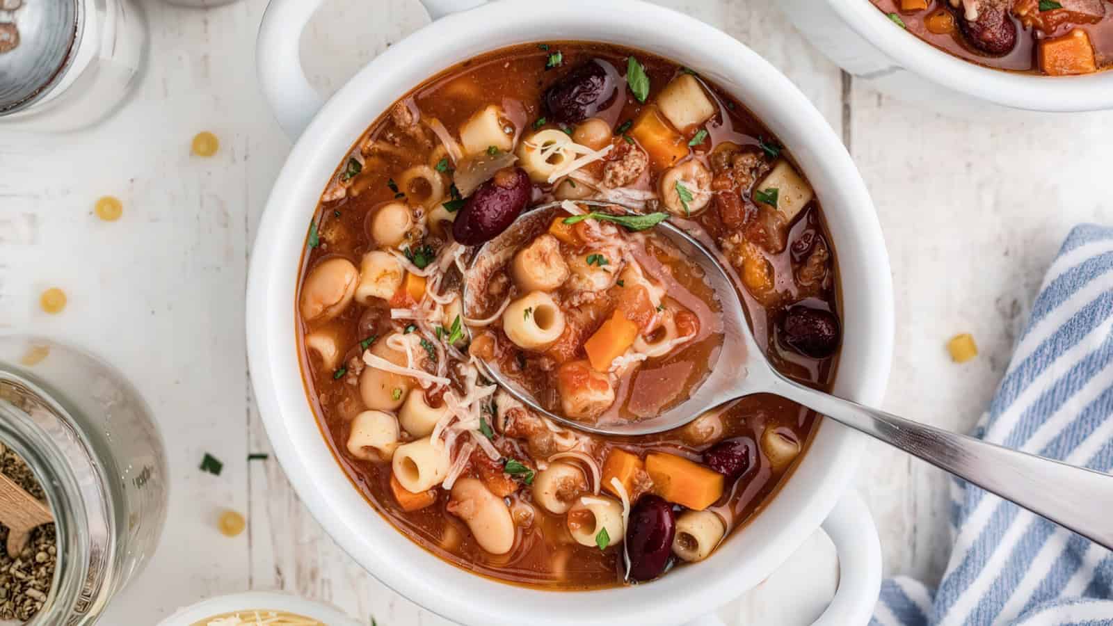 A bowl of minestrone soup with vegetables, pasta, beans, and herbs. A spoon rests inside, and the soup is garnished with shredded cheese. The background shows a striped napkin and part of a jar.