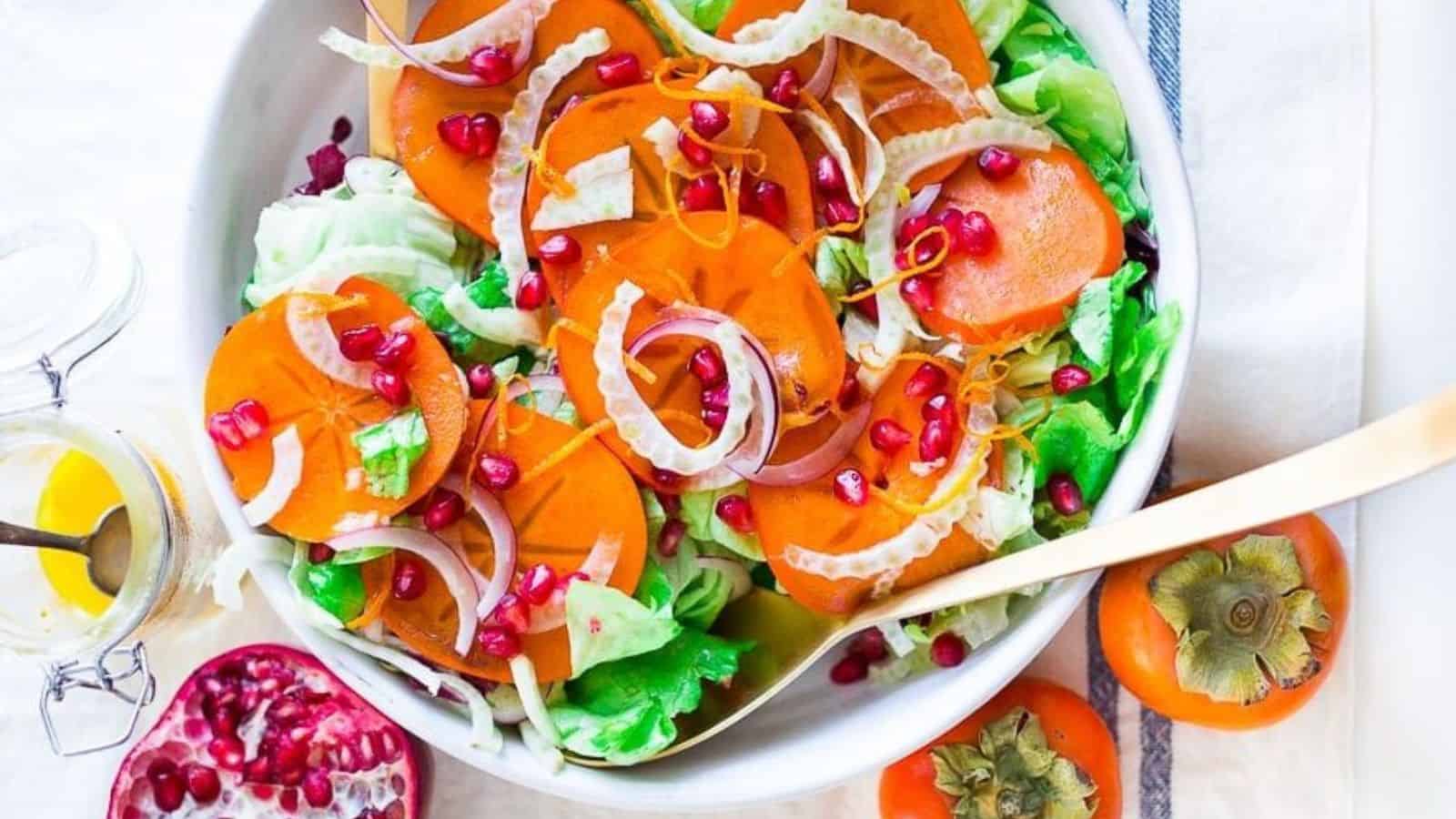 A bowl of vibrant salad featuring sliced persimmons, lettuce, fennel, red onion, and pomegranate seeds. A halved pomegranate and whole persimmons are next to the bowl, with a jar of dressing nearby.