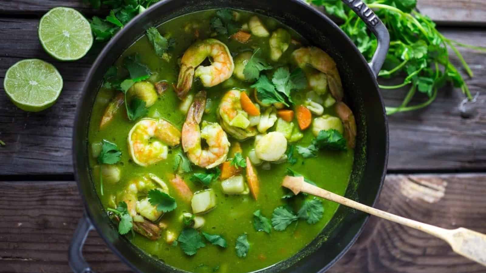 A pot of green seafood stew containing shrimp, scallops, and vegetables, garnished with fresh cilantro. A wooden spoon rests inside the pot, and lime halves with cilantro are nearby on a wooden table.