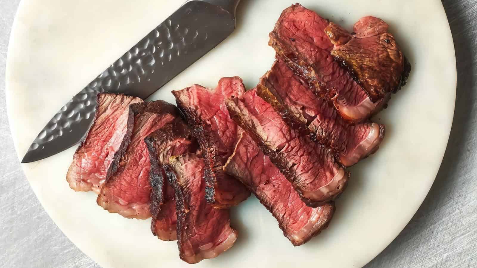 Sliced medium-rare steak with a seared crust is arranged on a white plate next to a textured knife, showcasing a juicy pink interior against dark brown edges.