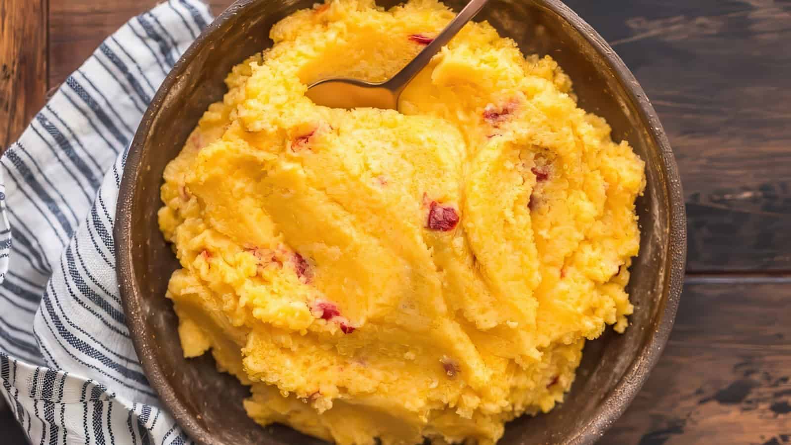 A bowl of creamy, cheesy pimento grits with visible pieces of pimento, served with a spoon. The bowl is placed on a wooden table next to a striped cloth.