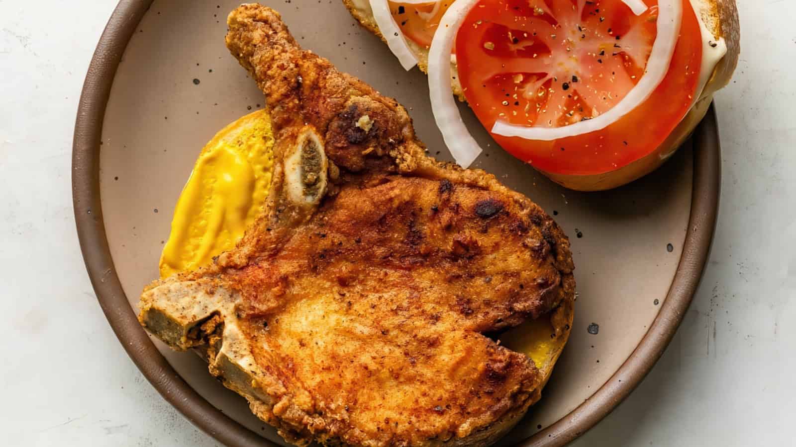 A plate with a fried pork chop resting on a slice of bread covered with mustard. Beside it, there are slices of tomato and onion on another slice of bread.