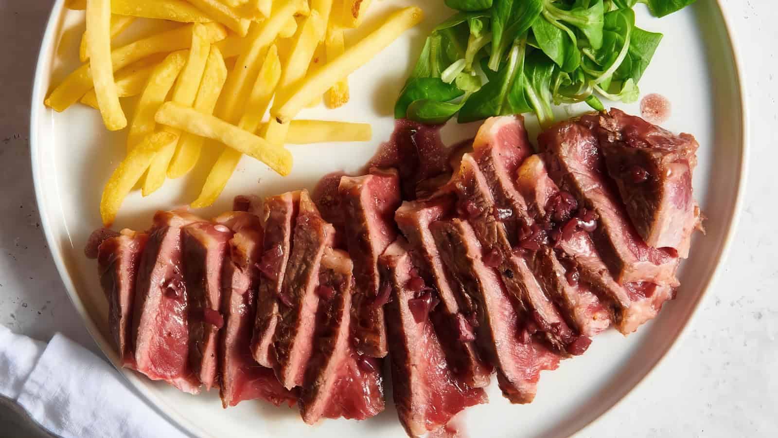 A plate of medium-rare sliced steak drizzled with a dark sauce, served with French fries and a green salad.