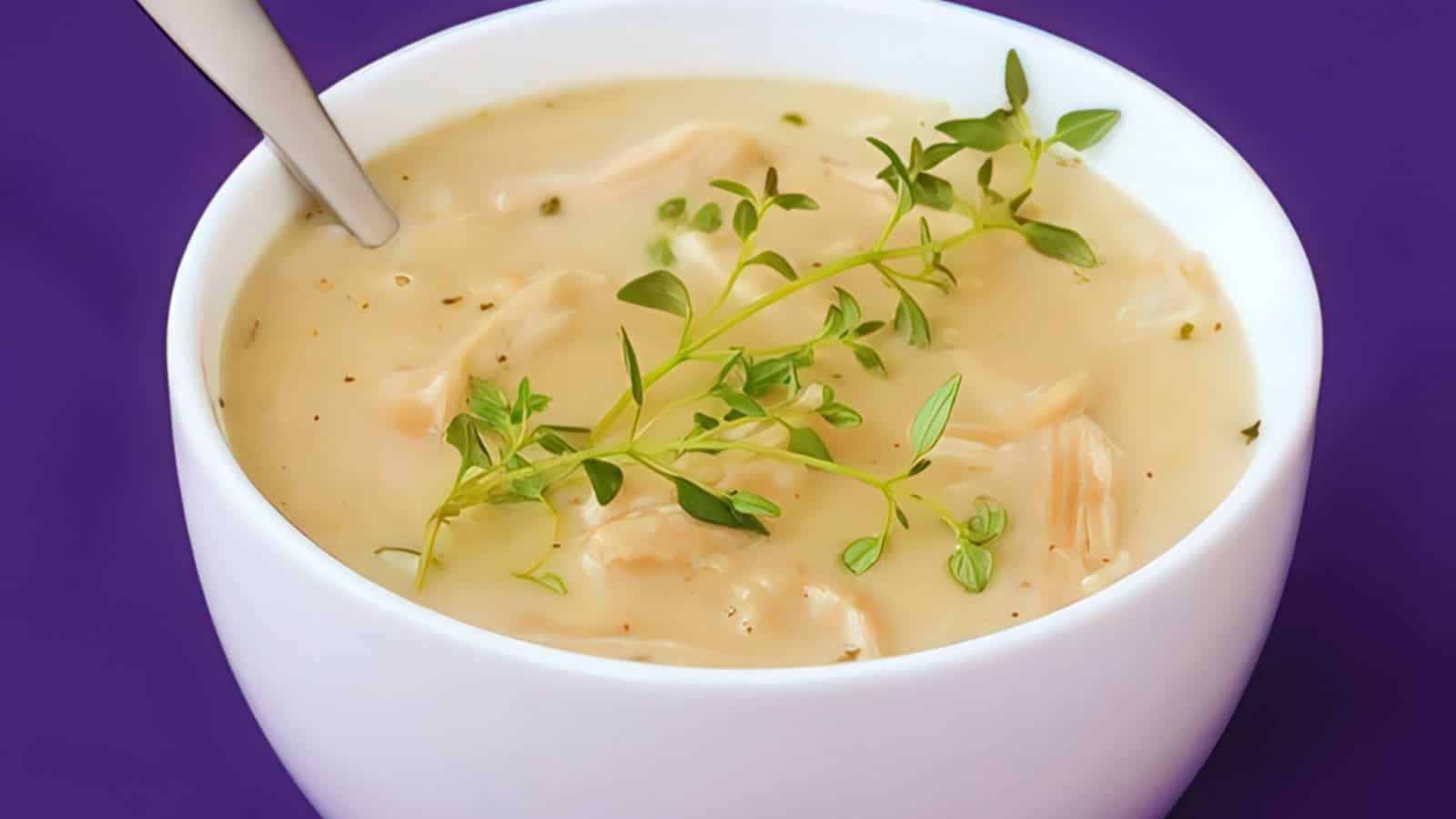 A bowl of creamy chicken soup garnished with fresh thyme. The soup contains shredded chicken pieces and is set against a rich purple background. A spoon is placed in the bowl, ready for serving.