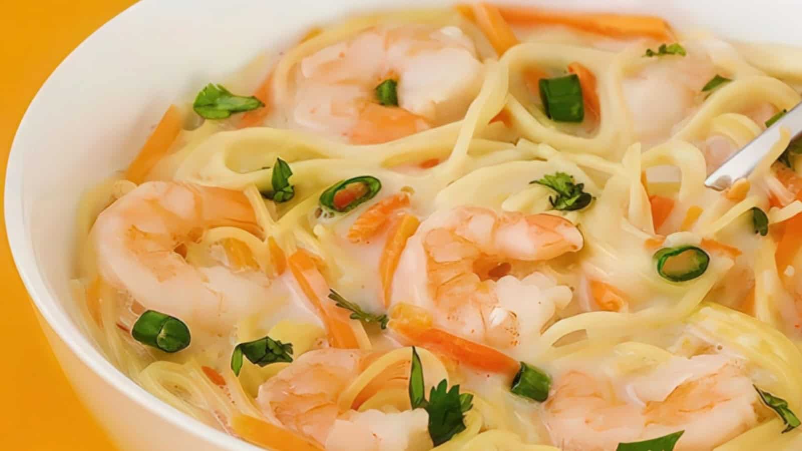 A bowl of creamy noodle soup topped with shrimp, sliced carrots, green onions, and fresh cilantro. The soup features a rich, milky broth and vibrant orange and green garnishes, creating a colorful and appetizing presentation.