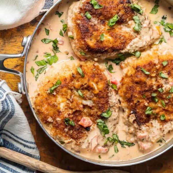 A skillet with three breaded chicken breasts in creamy sauce, garnished with chopped herbs, on a wooden surface next to a cloth napkin.