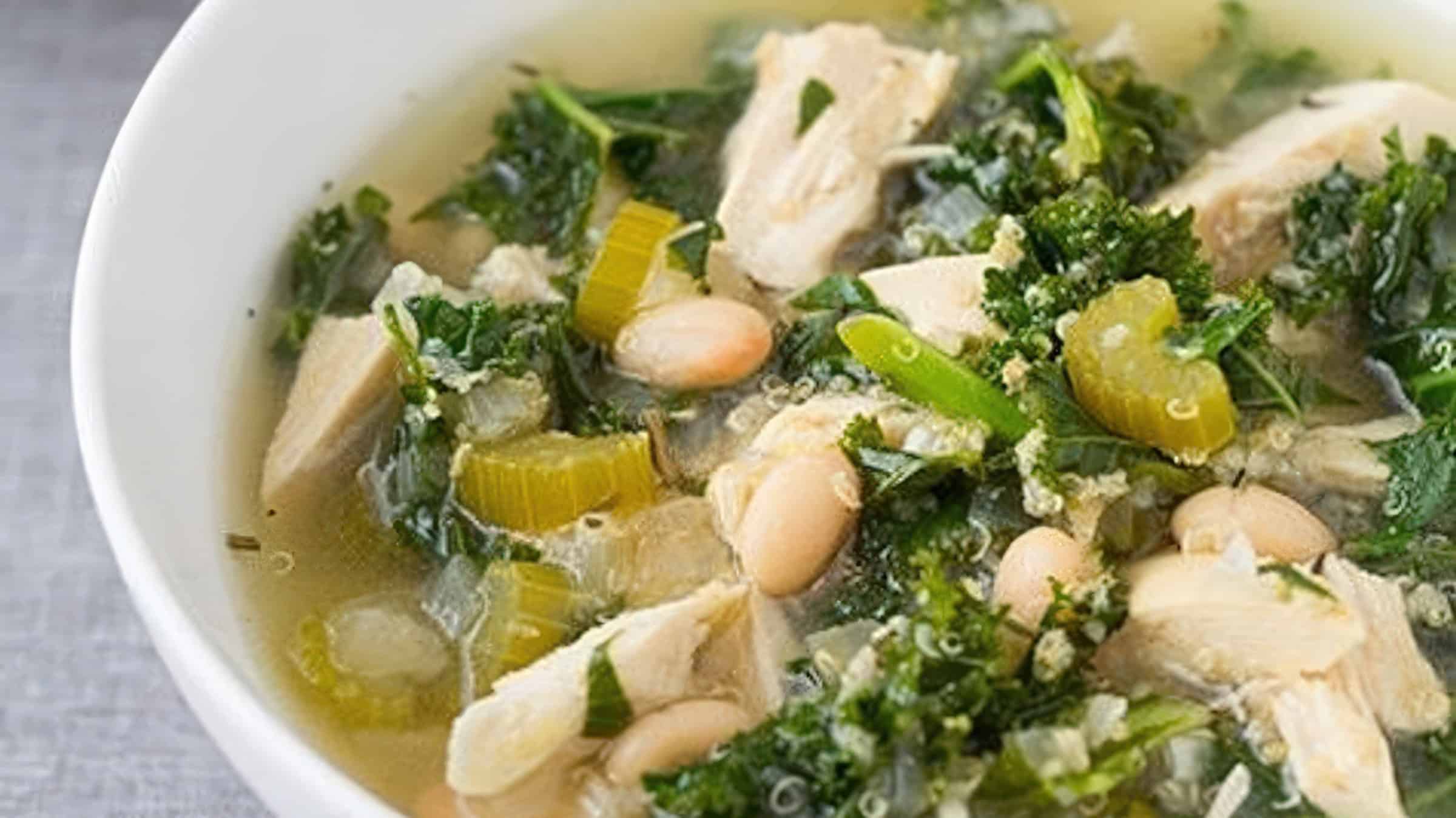 A bowl of soup with chunks of chicken, kale, celery slices, white beans, and herbs. The soup is clear and hearty, garnished with green vegetables and served in a white bowl on a gray surface.