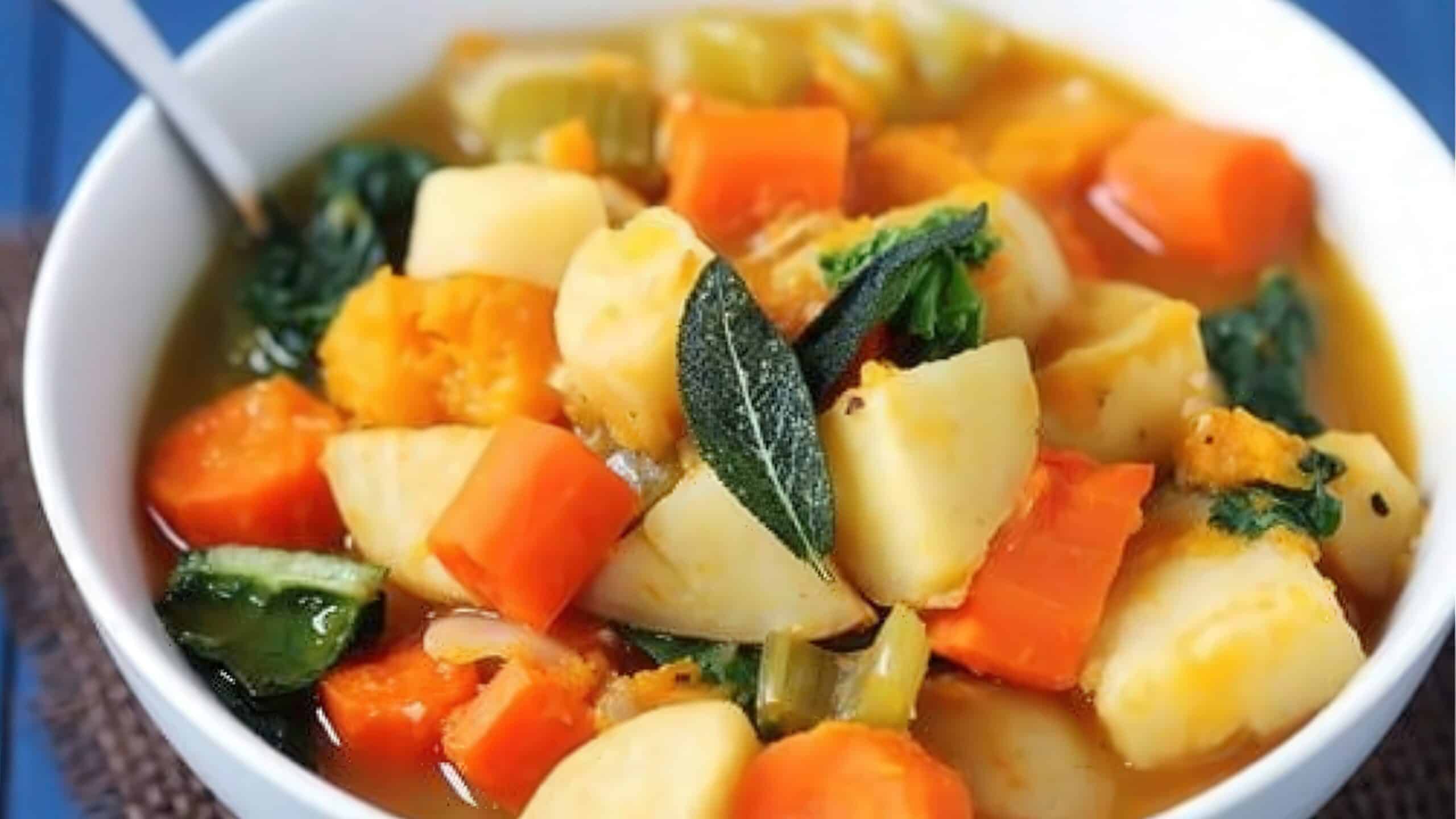 A bowl of hearty vegetable stew containing chunks of potatoes, carrots, and squash in a rich broth. Garnished with fresh sage leaves and served with a silver spoon.