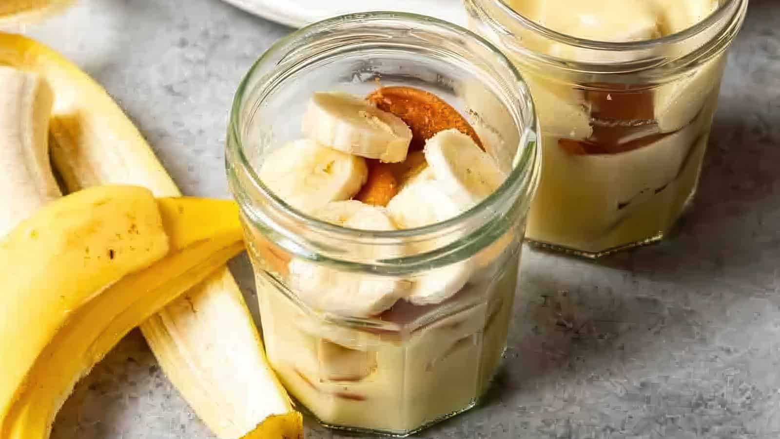 Two glass jars filled with pudding, sliced bananas, and vanilla wafers sit on a gray surface. A peeled banana is positioned on the left side of the image.