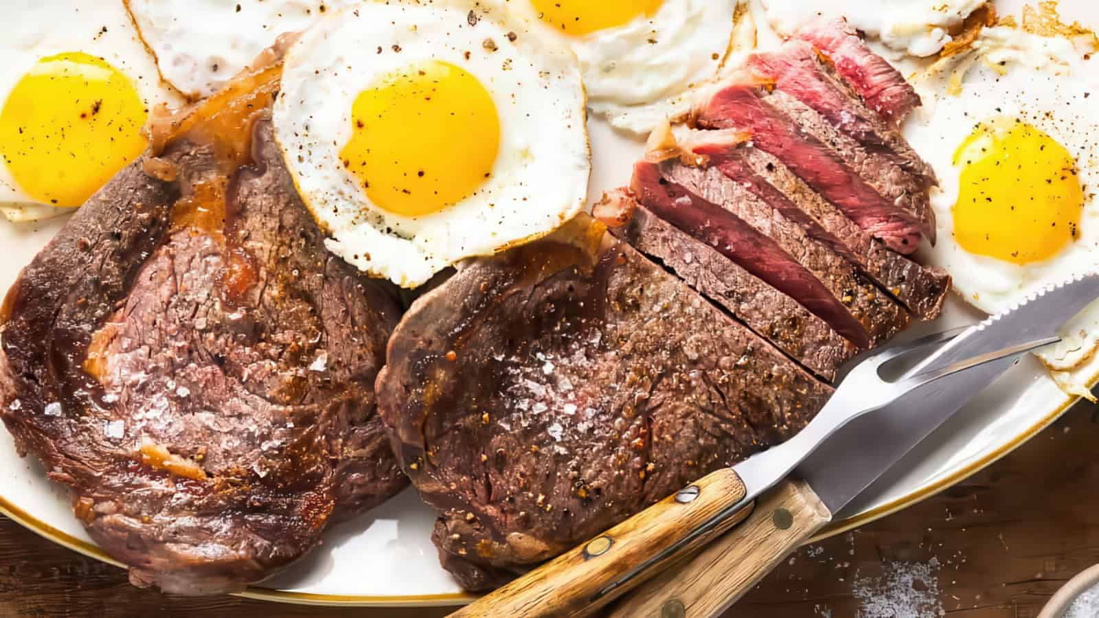 A plate with sliced steak, seasoned with salt, and four sunny-side-up eggs sprinkled with pepper. A knife and fork with wooden handles are placed on the right side of the plate.