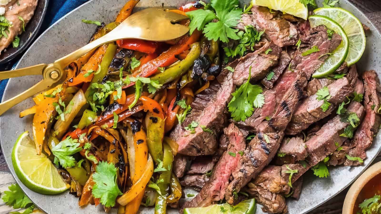 A plate filled with sliced grilled steak, garnished with lime wedges and cilantro, is accompanied by sautéed bell peppers and onions. Gold serving utensils rest on the side of the dish.