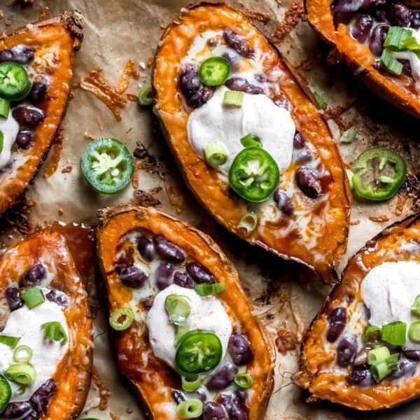 Baked sweet potato halves topped with melted cheese, black beans, sour cream, sliced jalapeños, and chopped green onions, arranged on a parchment-lined tray.