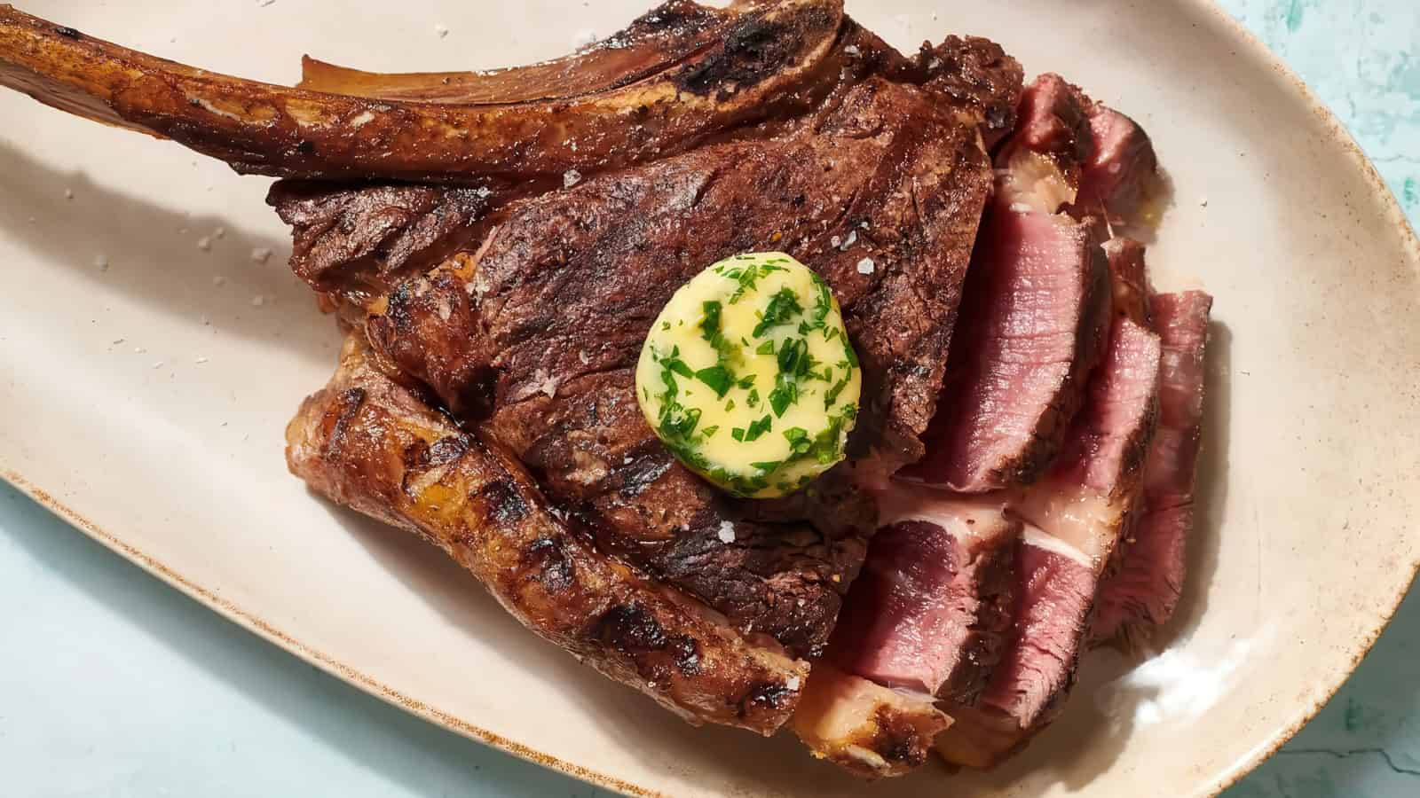 A juicy, grilled tomahawk steak is served on an oval platter. The steak is sliced and topped with a pat of herb-infused butter, creating a mouth-watering presentation.