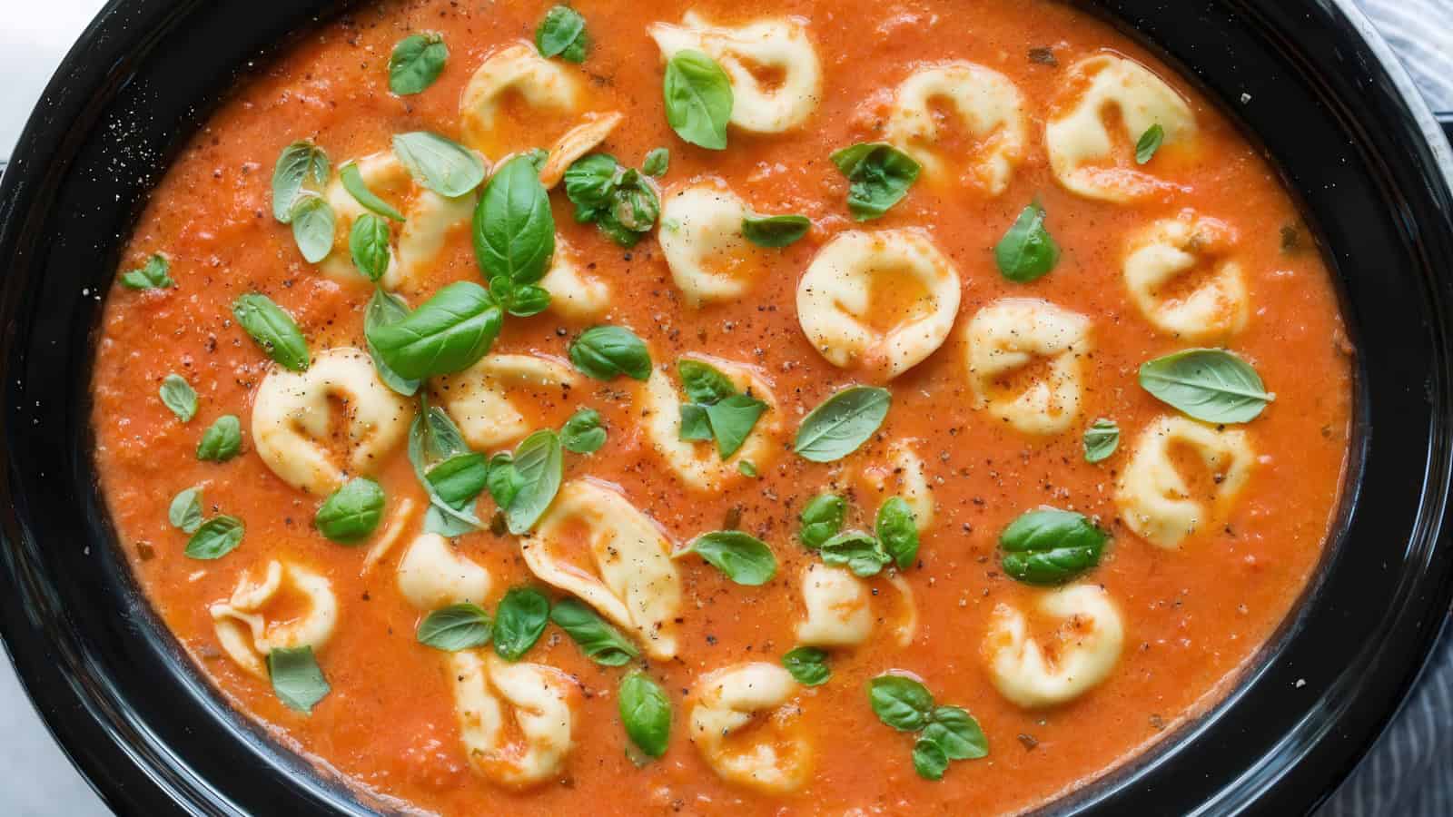 A bowl of creamy tomato tortellini soup garnished with fresh green basil leaves. The soup features a rich red tomato base, with scattered tortellini pieces sitting on the surface.