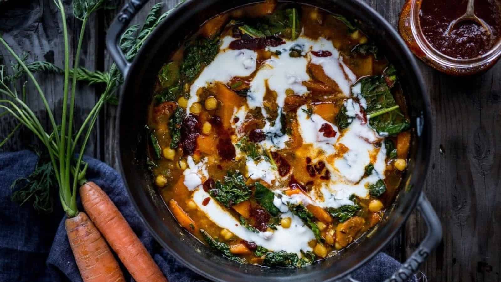A pot of vibrant stew containing chickpeas, carrots, greens, and spices, topped with a swirl of creamy yogurt. Fresh carrots are placed beside the pot on a dark wooden table, adding to the rustic presentation.
