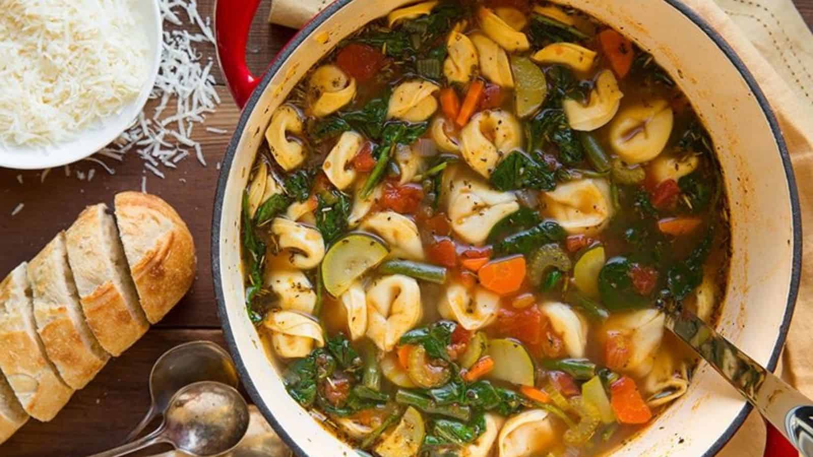 A pot of tortellini soup with spinach, zucchini, tomatoes, and green beans, served on a wooden table. Slices of bread and shredded cheese are on the side, along with two spoons.