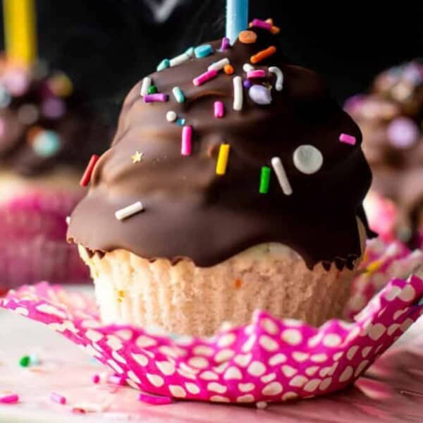 A cupcake with chocolate frosting and colorful sprinkles is placed in a pink and white polka dot wrapper. The background features two blurred cupcakes with similar decorations.