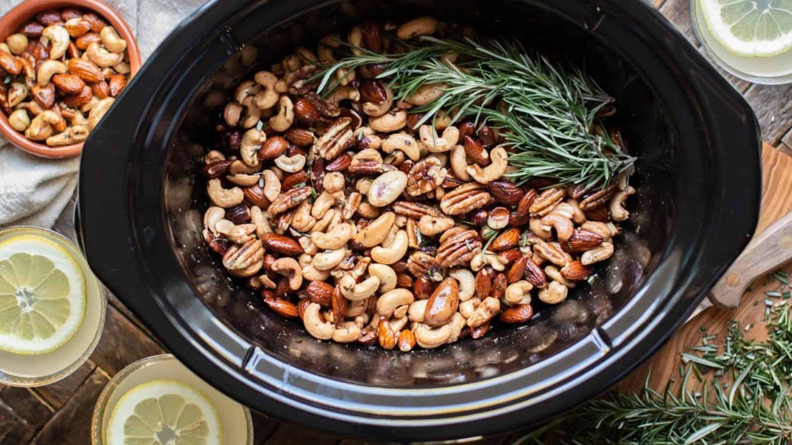 A black slow cooker filled with mixed nuts, including almonds, cashews, and pecans, garnished with fresh rosemary sprigs. Lemon slices and rosemary are placed nearby on a wooden surface.