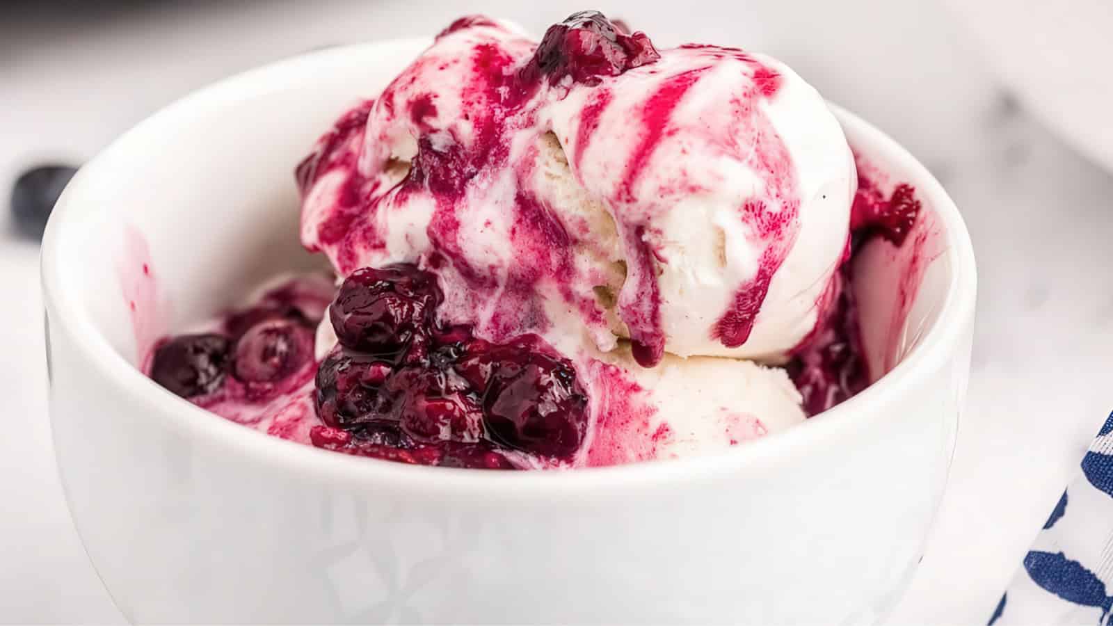 A white bowl filled with scoops of vanilla ice cream topped with rich, dark berry sauce, which is dripping over the sides. The dessert is served in a white, glossy bowl, creating a visually appealing contrast.