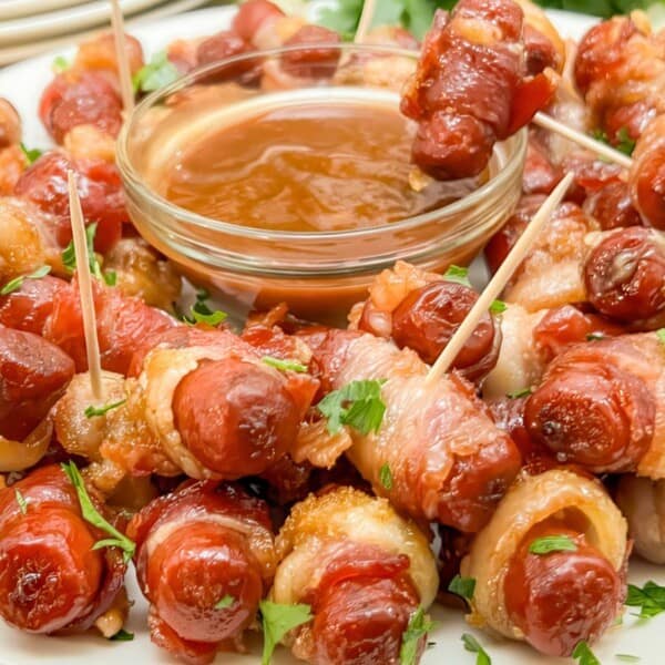 A plate filled with bacon-wrapped sausages on toothpicks, garnished with herbs. A small bowl of dipping sauce is in the center. The background shows a stack of plates.