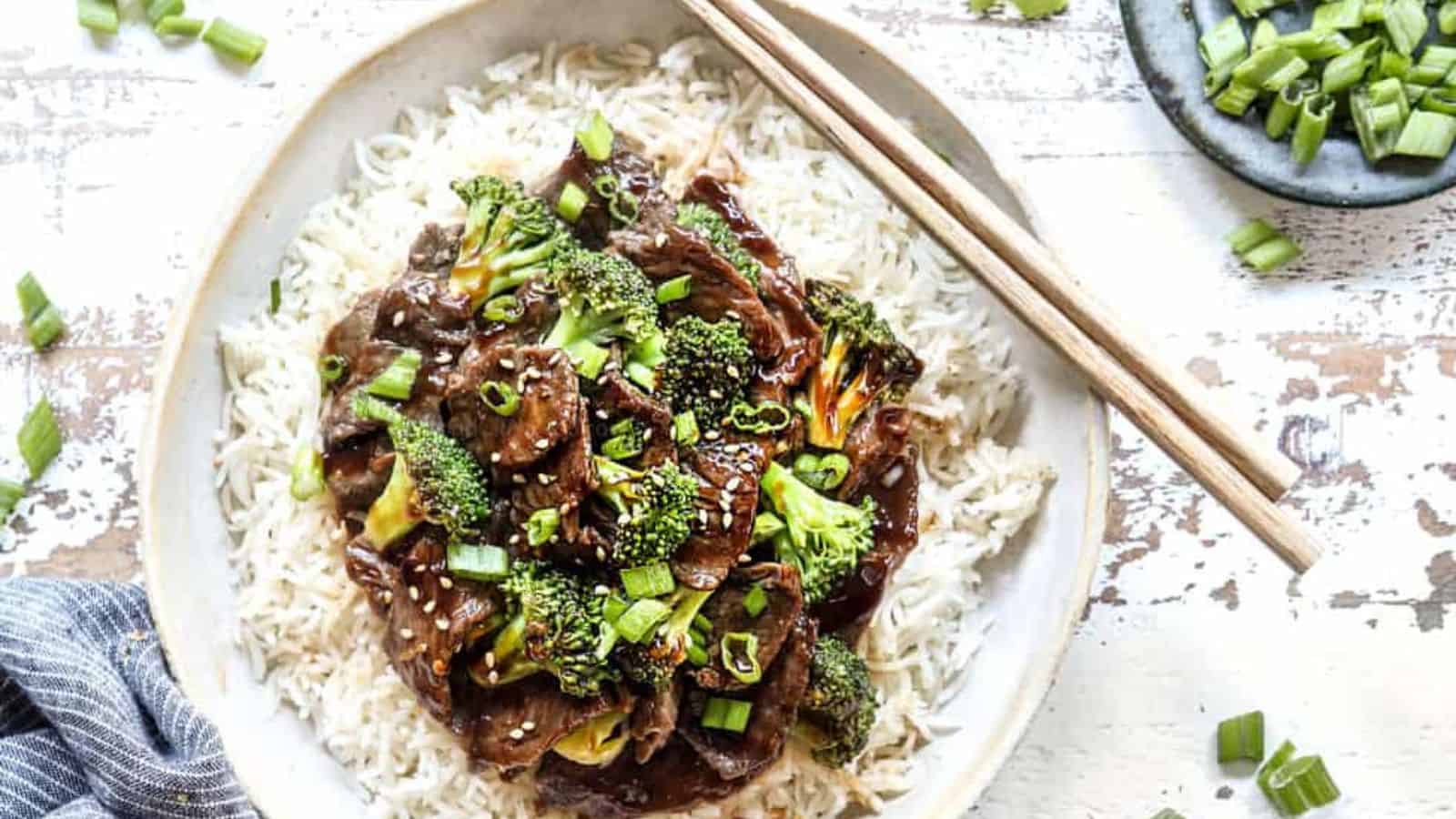 A plate of stir-fried beef and broccoli on white rice, garnished with green onions and sesame seeds, with chopsticks beside it.