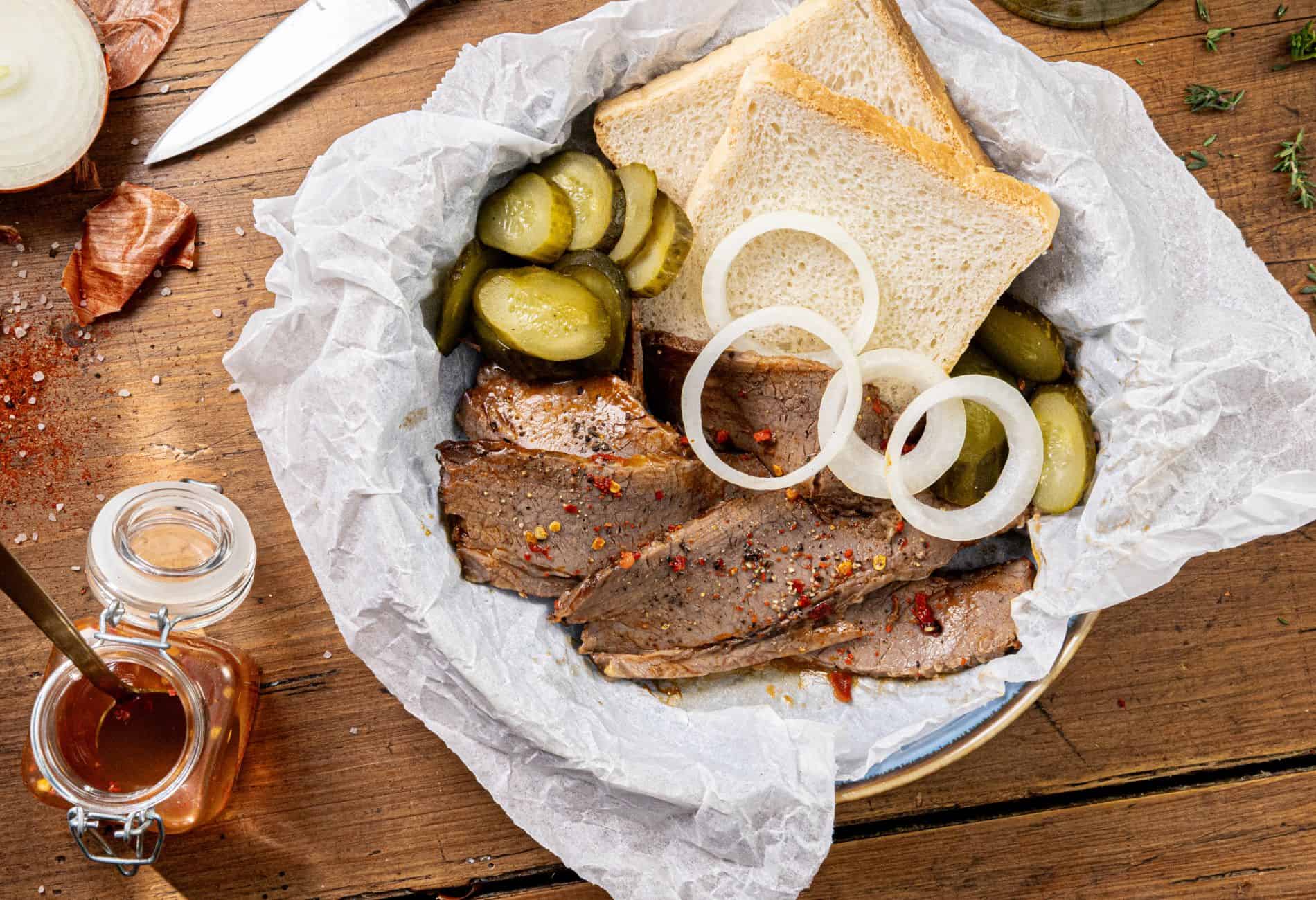 Slices of roasted beef topped with onion rings and accompanied by pickle slices and two pieces of white bread on a parchment-lined plate. A jar of sauce, knife, onion, and herbs are on the wooden table surface.