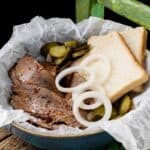 A bowl with beer-braised beef brisket, pickle slices, onion rings, and two pieces of white bread sits on parchment paper.