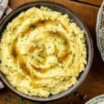 A bowl of browned butter mashed potatoes garnished with herbs and drizzled with melted butter rests on a wooden table, beside a patterned plate and a plaid napkin.