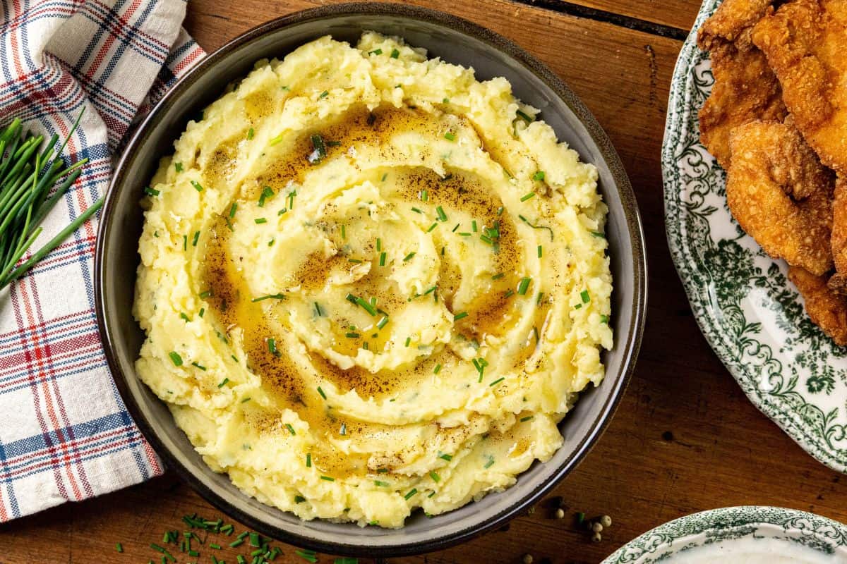 A bowl of browned butter mashed potatoes garnished with herbs and drizzled with melted butter rests on a wooden table, beside a patterned plate and a plaid napkin.