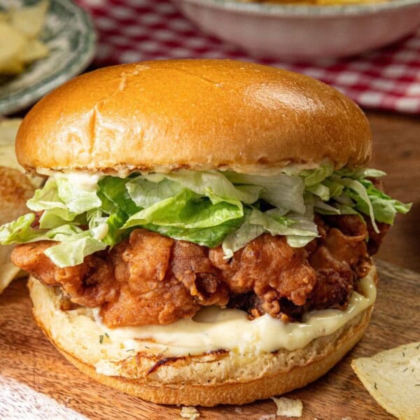 The image shows a crispy chicken sandwich with lettuce and mayonnaise in a soft bun, accompanied by potato chips on a wooden board.