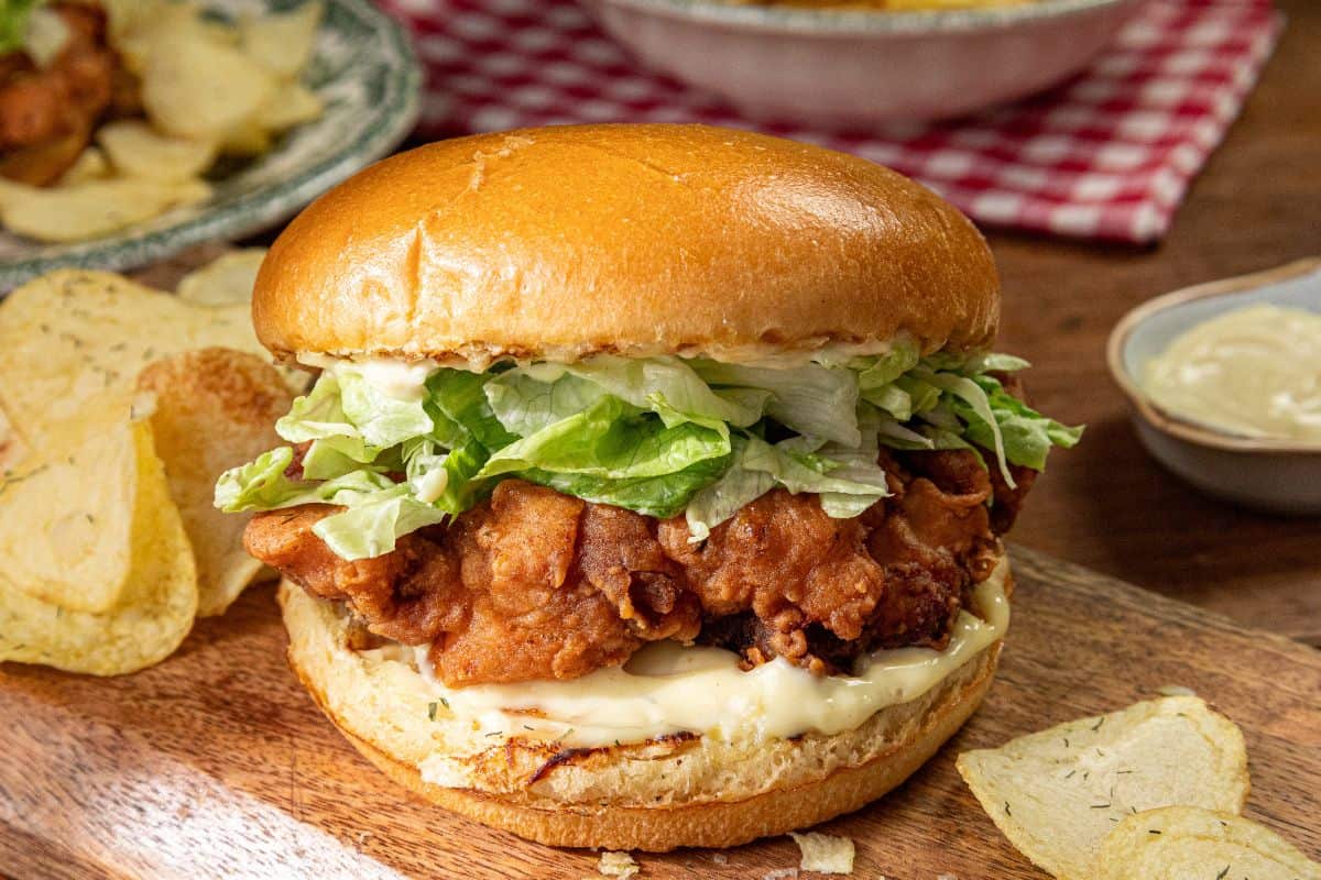 The image shows a crispy chicken sandwich with lettuce and mayonnaise in a soft bun, accompanied by potato chips on a wooden board.
