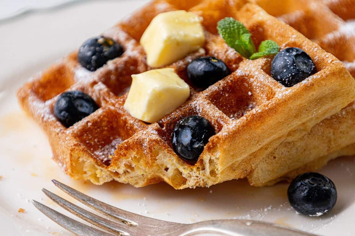 A stack of cornmeal waffles topped with blueberries, a pat of butter, a mint leaf, and a dusting of powdered sugar sits on a plate next to a fork.