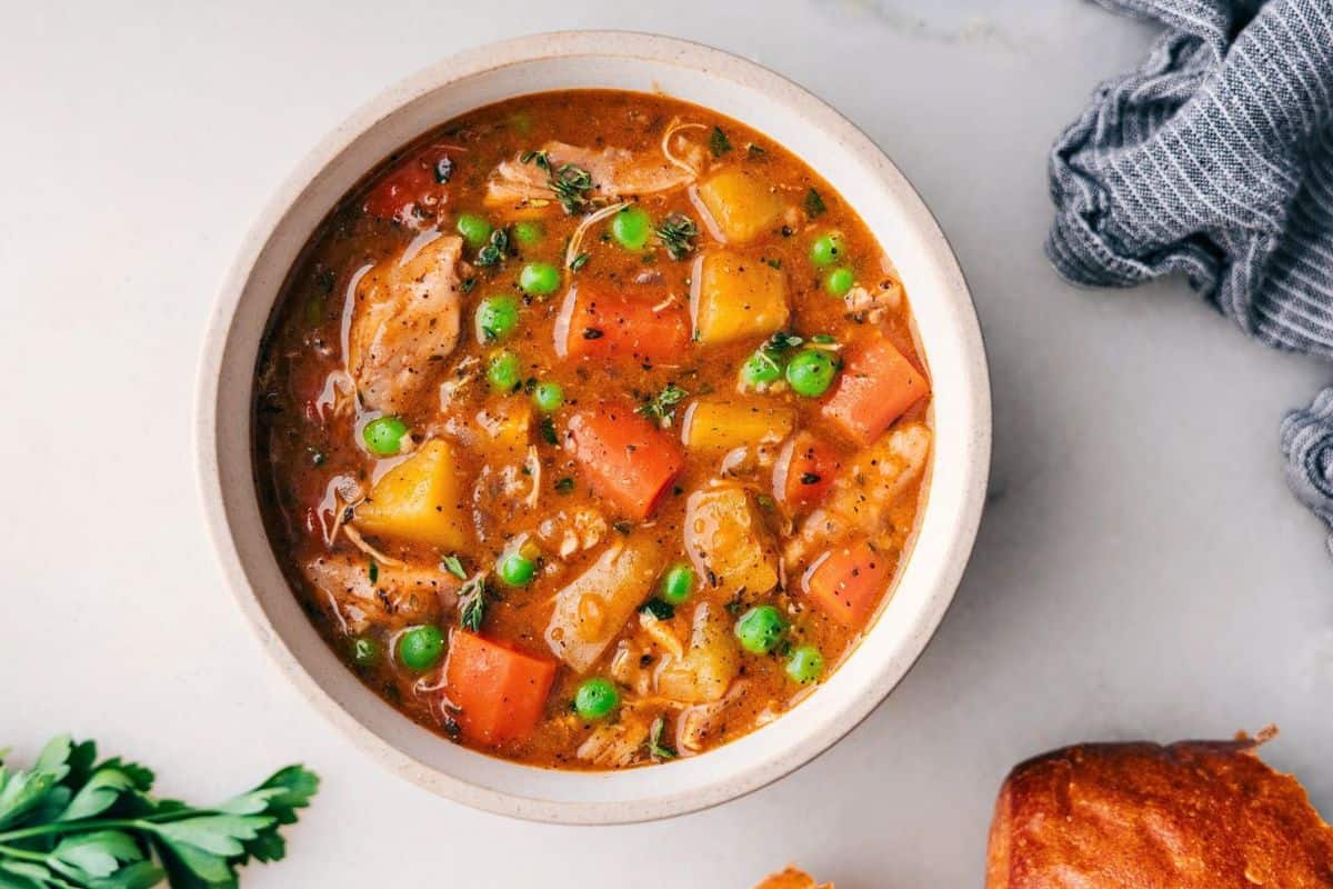 A bowl of hearty vegetable stew featuring chunks of carrots, potatoes, peas, and herbs in a rich broth. A piece of bread is partially visible on the side, and a gray napkin is nearby on a light surface.