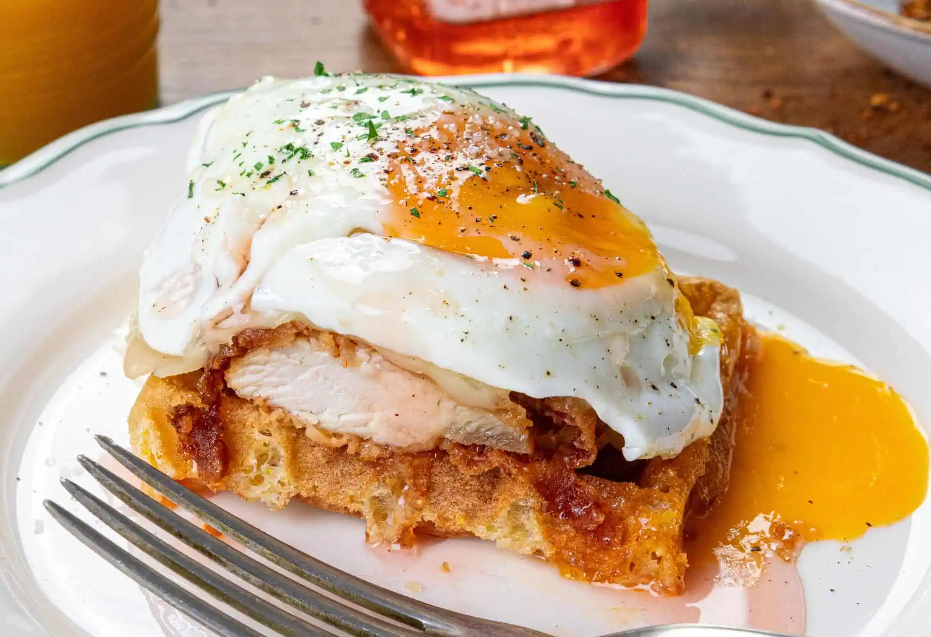 A crispy waffle topped with fried chicken and a poached egg, with a runny yolk and sprinkle of herbs on a white plate.